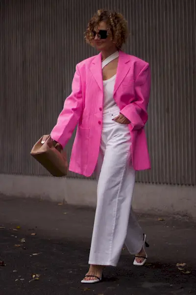 A woman wears a pink blazer, pink top, pink jeans, beige clutch, and open toe sandals.