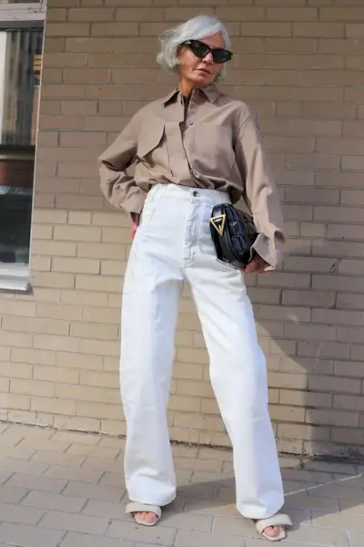 A woman wears a brown button-down shirt, black bottega veneta bag, white wide- leg pants, and beige sandals.