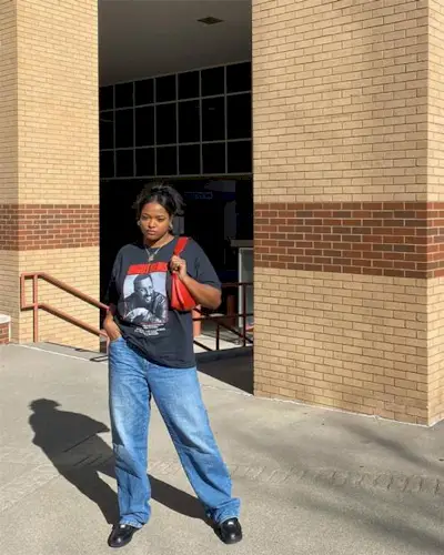 A woman wears a graphic t-shirt, a red bag, blue jeans, and black loafers.