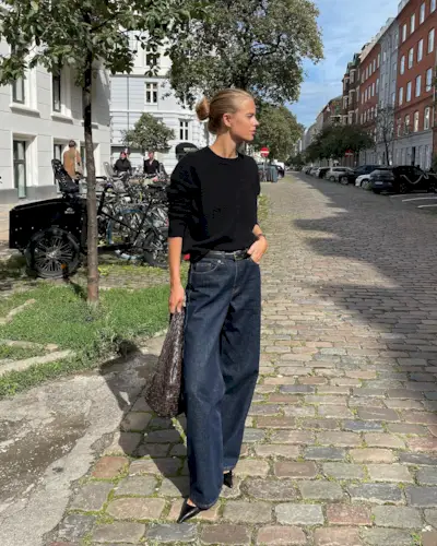 Clara wears a black sweater, wide-leg jeans, black pointed toe heels, and a brown bag.