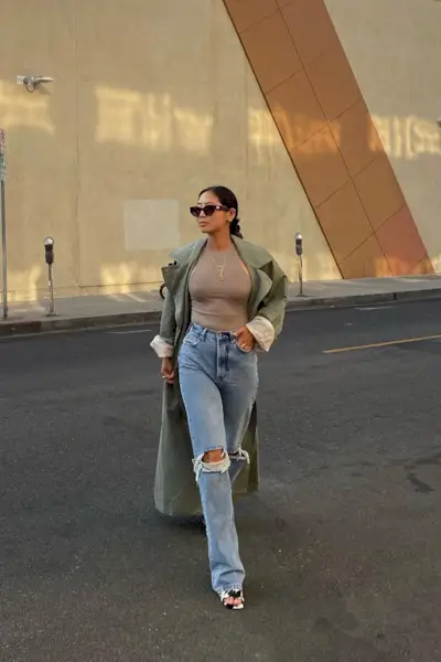 a photo showing how to wear wide jeans with a woman wearing a green trench coat over a tan tank top and wide leg jeans