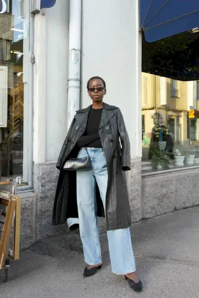 Sylvie wears a leather trench coat, black long-sleeve, wide-leg jeans, black kitten heels, and a black bag.