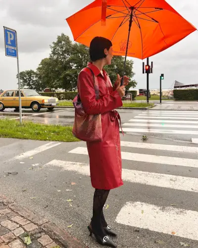 A woman wears a red leather trench coat, tights, black slingbacks, and a sequin shoulder bag.