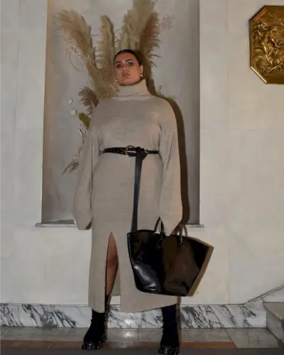 Isabelle wears a turtleneck sweater dress in beige with a black belt, black boots, and a black shiny tote bag.