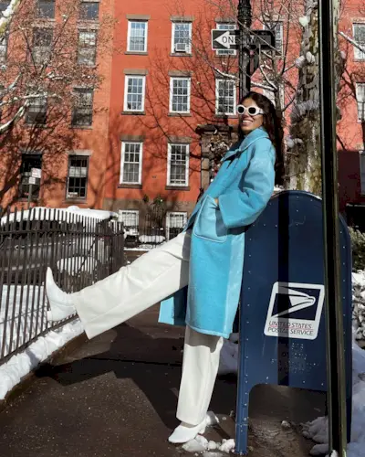 A woman wears a long blue coat, white straight-leg jeans, white boots, and white sunglasses.