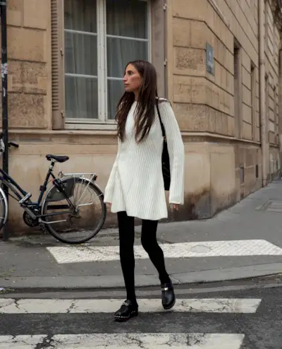 A woman wears a white sweater dress, black tights, and black clogs.