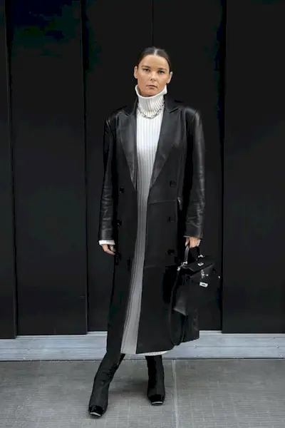 a photo showing how to wear dresses in the winter with a woman wearing a black long leather coat over a white turtleneck dress with black knee-high boots