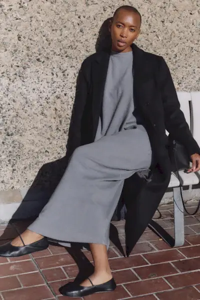 a photo showing how to wear dresses in the winter with a woman wearing a long black wool coat over a gray knit maxi dress and black ballet flats