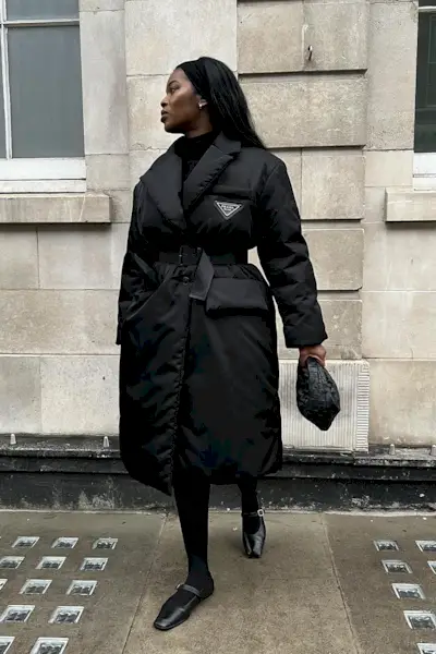 a photo showing how to wear dresses in the winter with a woman wearing a black puffer coat over a sweater dress with sheer tights and black flat mary janes