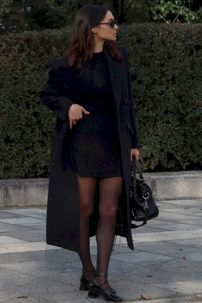 a photo showing how to wear dresses in the winter with a woman wearing a long navy coat over a black mini dress with sheer black tights and black mary jane flats