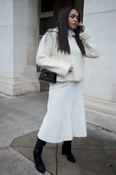 a photo showing how to wear dresses in the winter with a woman wearing a white turtleneck dress under a matching zip-up jumper styled with black knee high boots and a black shoulder bag