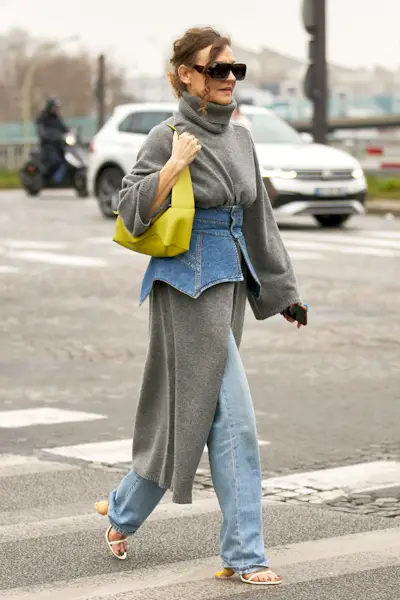 a photo showing how to wear dresses in the winter with a woman wearing a gray sweaterdress over blue jeans with strappy sandals and a corset belt