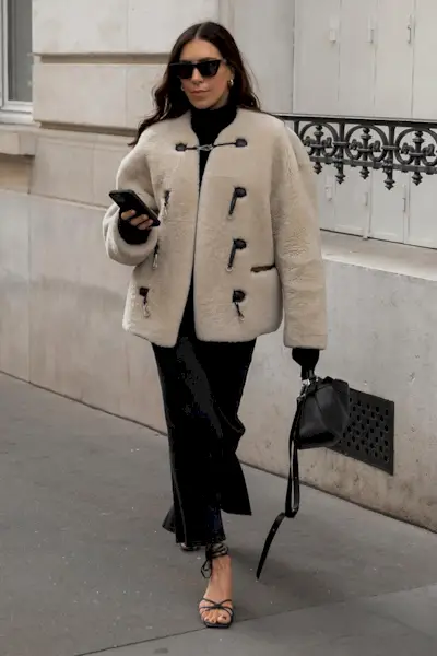 a photo showing how to wear dresses in the winter with a woman wearing a black turtleneck dress layered under a tan shearling jacket styled with black sandals