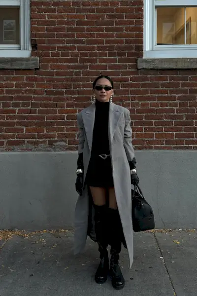 a photo showing how to wear dresses in the winter with a woman wearing. a black turtleneck dress under a long gray coat with black over-the-knee boots and silver jewelry