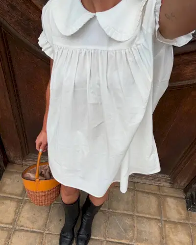 A woman wearing a white collared dress, black tall boots, and a wicker bucket bag.