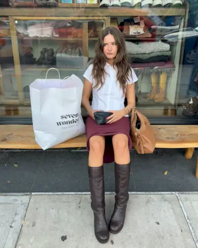 A woman sitting down wearing a red skirt, a white blouse, and brown leather tall boots.