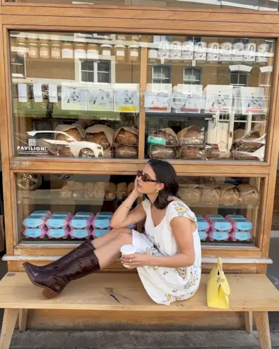 A woman sitting down on a bench wearing brown leather cowgirl boots, a floral print dress, and sunglasses.