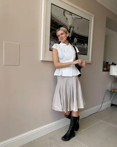 A woman standing wearing a white collared blouse, a pleated skirt, and black leather motor boots.