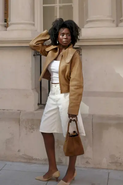 How to style white jeans for fall, shown in a photo of a woman standing outside on the sidewalk wearing silver earrings with a tan leather cropped jacket styled with a white tank top, white long jean shorts, tan suede pumps, and a tan bucket bag