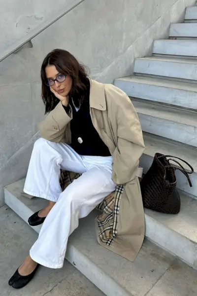 how to style white jeans for fall shown in a photo of a woman sitting outside on concrete stairs wearing a tan trench coat over a black crewneck sweater styled with white wide-leg jeans, black tabi ballet flats, a long necklace, black eyeglasses, and a black handbag