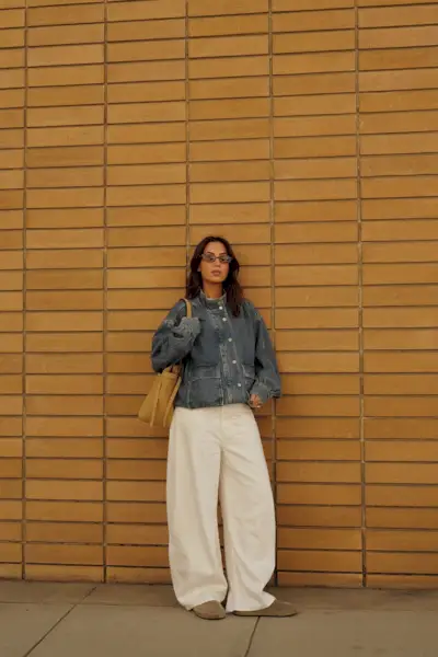 How to style white jeans for fall, shown in a photo of a woman standing outside against a yellow brick wall, wearing brown sunglasses, an oversize denim funnel neck jacket, a white t-shirt, white wide-leg jeans, suede Birkenstocks, and an oversize brown tote bag