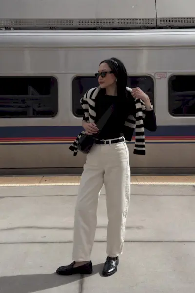 How to style white jeans for fall, shown in a photo of a woman standing outside at a train station wearing a black headband, black sunglasses, a black boatneck top with a striped sweater over the shoulders, a black belt, and white baggy jeans, and black loafers