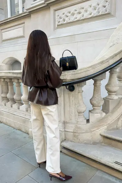 How to style white jeans for fall, shown in a photo of a woman standing outside near a white marble staircase wearing a burgundy leather cropped jacket with white straight-leg jeans, burgundy slingback heels, and a black structured bag