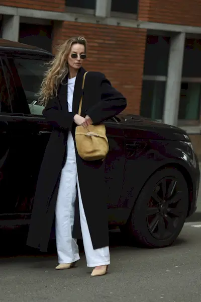 How to style white jeans for fall, shown in a photo of a woman standing outside next to a black car wearing a long black coat over a white t-shirt styled with white relaxed jeans, tan suede pumps, a tan suede bucket bag, and gold wireframe sunglasses