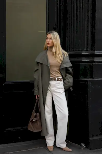 How to style white jeans for fall, shown in a photo of a woman standing outside against a black building wearing a green trench coat over a tan turtleneck with a tan studded belt, white wide-leg jeans, tan suede ankle boots, and a brown bowler bag