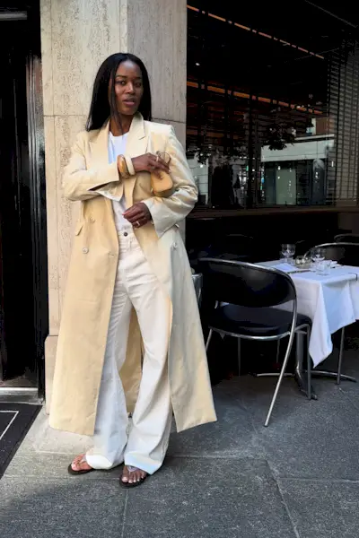 how to style white jeans for fall shown in a photo of a woman standing outside a restaurant wearing an off-white long coat over a white t-shirt styled with tan bangles, a brown-butter colored bag, white baggy wide-leg jeans, and black jelly flip flops