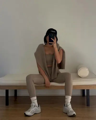 Jordan wears beige leggings, a beige sports bra, beige sweater, black hat, white tube socks, and gray new balance sneakers.
