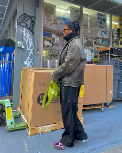 A woman wears a brown distressed leather jacket, black turtleneck, black cargo pants, burgundy new balance sneakers, and a green bag.