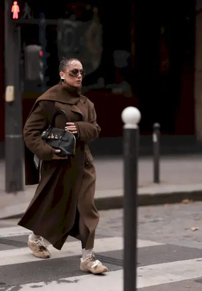 Anna Wein is seen wearing rimless oversized sunglasses with brown lenses, slim gold metal temples, and a gold metal nose bridge from Miu Miu; chunky squared earrings in gold with a black stone; an ankle-length brown wool trench coat featuring a double-button placket and notch lapels from Sandro; paired with a color-matching wide long scarf, also from Sandro; a rectangular black leather bag with gold details and several bag charms from Miu Miu; beige socks; and beige suede sneakers with fluffy lining and two different shoelaces in cream white cotton and brown leather from Miu Miu x New Balance during Paris Fashion Week on September 29, 2024 in Paris, France.