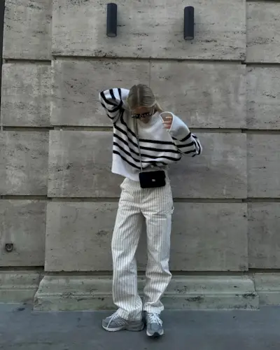 A woman wears a black and white stripe sweater, white stripe pants, and gray new balance sneakers.