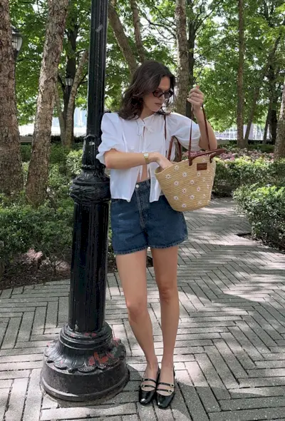 a woman's outfit showing what to wear in 80-degree weather with a white puff sleeve blouse styled with denim shorts and black mary jane ballet flats and a woven raffia tote bag
