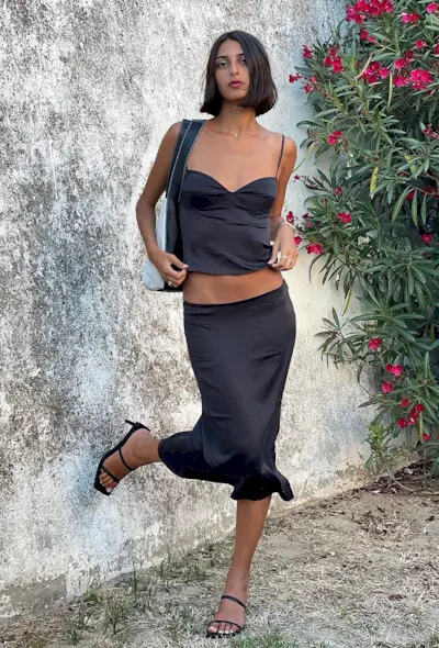 a woman's outfit showing what to wear in 80-degree weather with a black slip midi skirt styled with a matching black silk tank top, black strappy mule sandals, and a shoulder bag