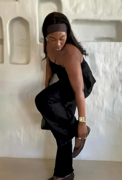 a woman's outfit showing what to wear in 80-degree weather with a black tube top styled with black trousers, brown ballet flats, gold jewelry, and a brown stretchy headband