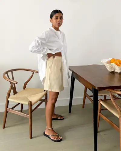 British style influencer Monikh Dale poses in her chic London dining room with an abstract white bowl full of oranges on the table wearing a white oversize line button-down shirt, butter-yellow shorts, anklets and black flat flip-flop sandals