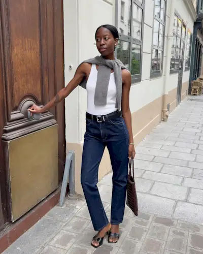 European fashion influencer Sylvie Mus poses on a Paris sidewalk wearing a gray sweater around her shoulders, a white tank top, black belt, dark-wash straight-leg jeans, a brown woven bag, and black square-tow mule heeled sandals
