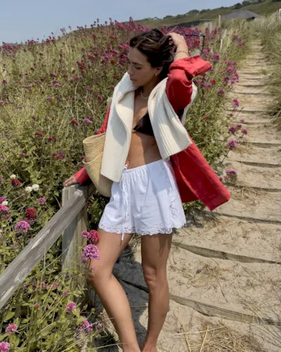 British style influencer Jessica Skye poses on a flower-lined beach path in Sennen Cove, Cornwall in the UK wearing a cream sweater over the shoulders, a red linen button-down shirt, black bikini top, straw tote bag, and lace-trimmed white cotton shorts.