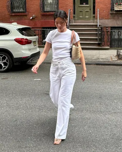 style influencer Mimi Nguyen walks across a New York City street wearing a small straw tote bag with brown leather straps, a fitted white T-shirt, white high-waisted straight-leg jeans, and neutral strappy heeled sandals