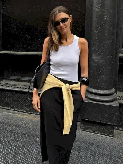 NYC style influencer Coco Schiffer poses on a sidewalk wearing black rectangular sunglasses, a white tank top, black minimal shoulder bag, a large black cuff bangle bracelet, yellow sweater tied around the waist, and a black midi skirt.