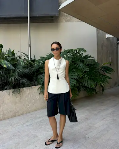 Stylist Debora wears the Reformation Jessie thong Sandal in black leather with black Bermuda shorts, a white scooped neck of the shoulder top a long black frayed pendent and 90s style oval sunglasses. she is carrying a black suede bag and is stood in front of some green palm trees in an image taken from her Instagram recently.