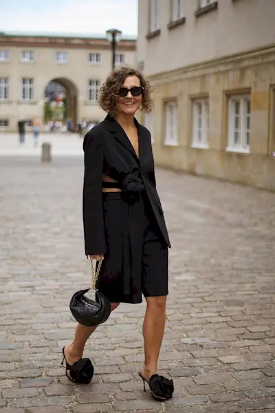 a photo showcasing one of the best summer work outfits with a woman wearing a black blazer with black bermuda shorts, black sandals, and a black crescent handbag