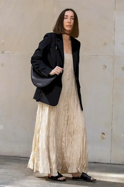 a photo showcasing one of the best summer work outfits with a woman wearing a black blazer over a tan plissé maxi dress with black slide sandals