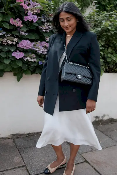 a photo showcasing one of the best summer work outfits with a woman wearing a black blazer over a white slip dress with two-tone ballet flats and a black quilted bag