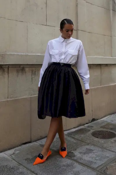 a photo showcasing one of the best summer work outfits with a woman wearing a white button-down shirt, a black full skirt, and orange slingback heels