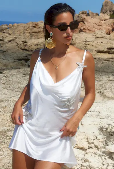 Shell jewelry trend shown on a woman wearing a white slip dress with oversize gold shell earrings and black sunglasses.