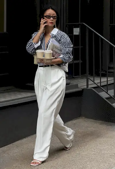 a photo showing what shoes to wear with wide-leg pants with a woman walking on the sidewalk wearing a blue and white striped button-down shirt tucked into white wide leg pants styled with white strappy heeled sandals