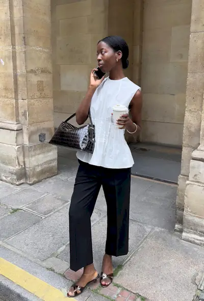 a photo showing what shoes to wear with wide-leg pants with a woman on the sidewalk wearing a white blouse with wide-leg cropped black trousers, brown open-toed mule sandals, and a brown handbag